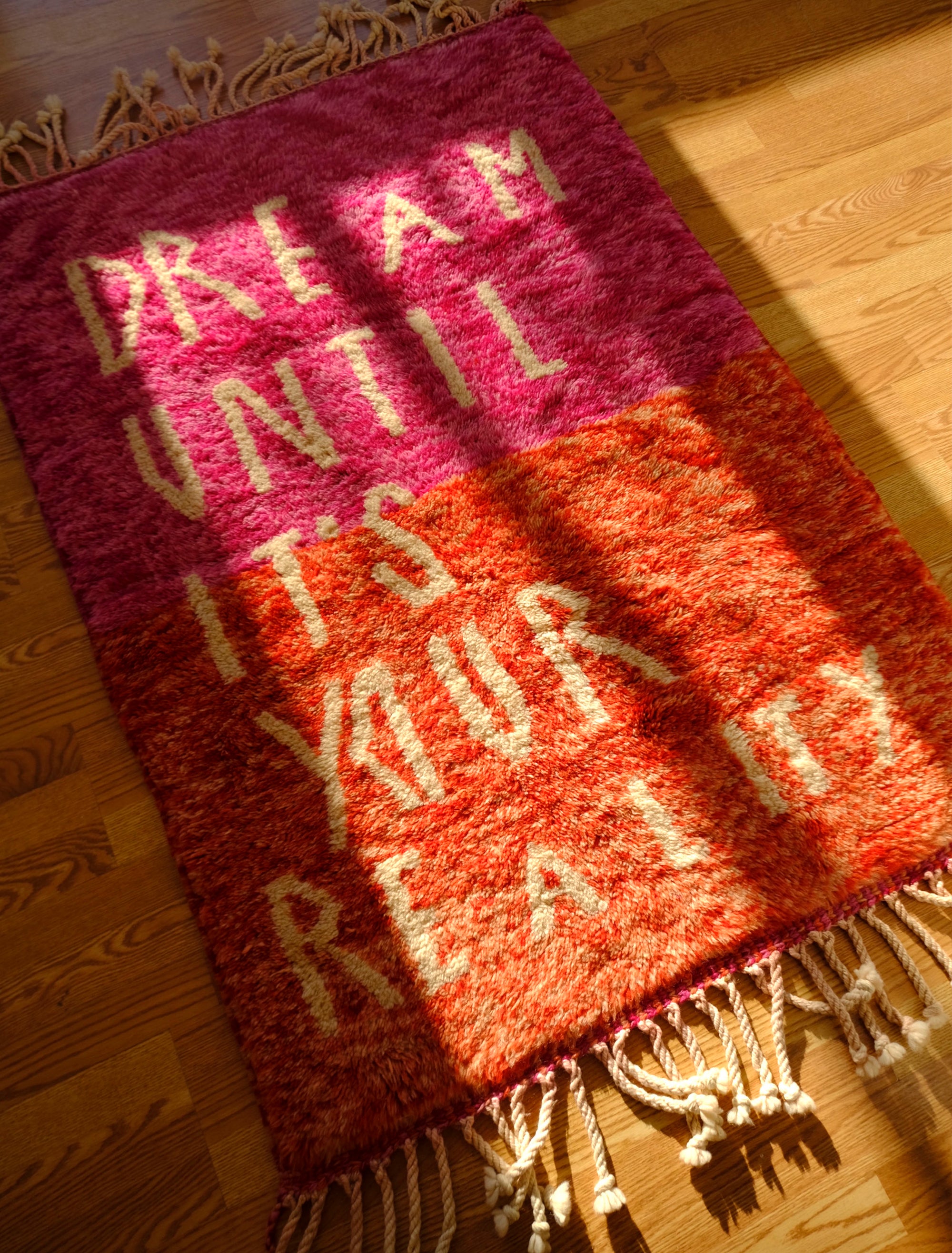 Introducing the enchanting 'Rosy Dreamscape Rug,' a delightful fusion of pink and red yarns forming a playful base, with the inspiring phrase 'Dream Until It Is Your Reality' expertly woven in white yarn. Elevate your space with this captivating blend of colors and motivational message, creating a joyful and imaginative ambiance that adds a touch of beauty and positivity to your home.